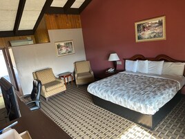 Un cuarto de hotel con una cama grande, dos sillones, una mesita, una mesita de noche con una lámpara y un cuadro en la pared.