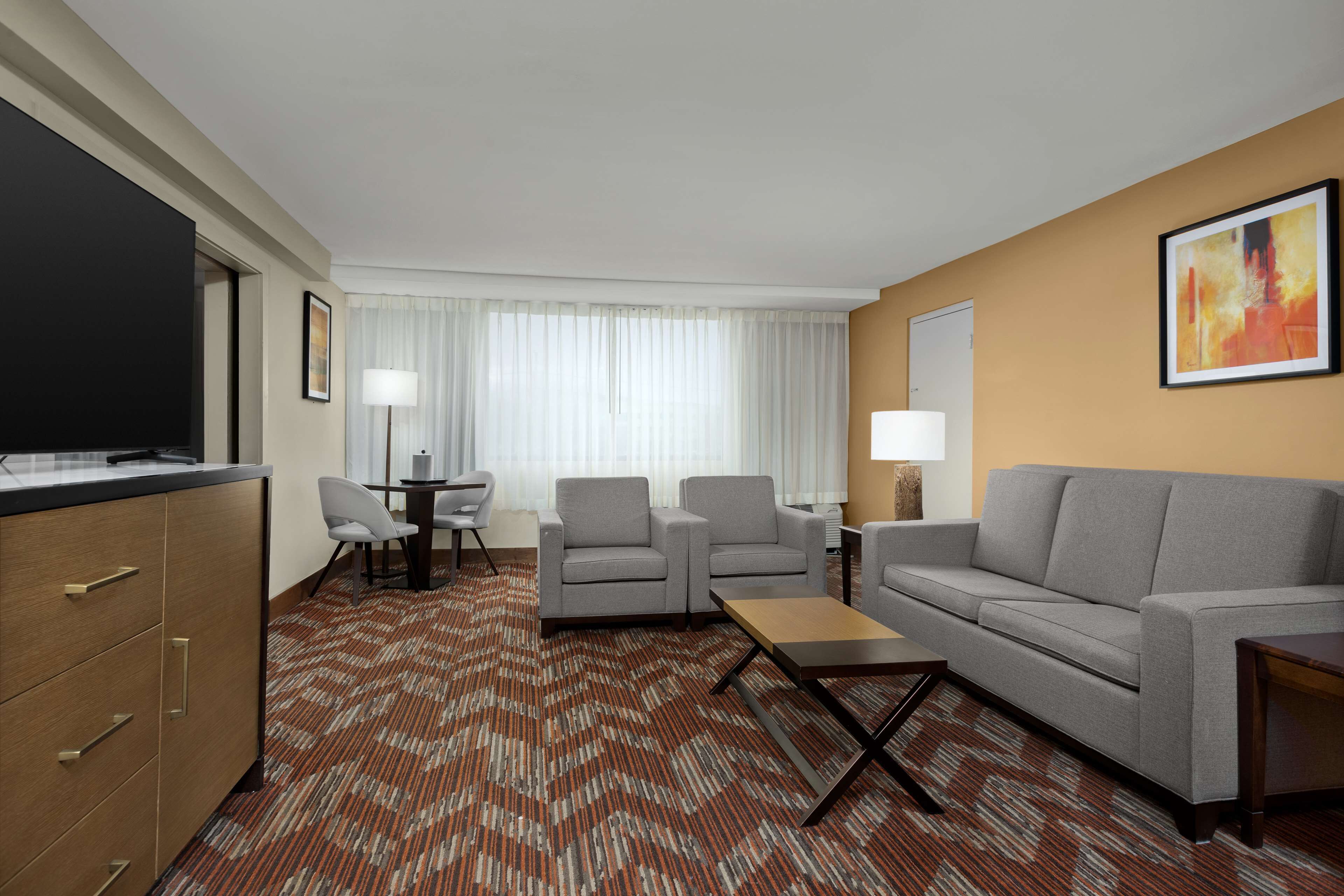 Quarto de hotel com sofá, duas poltronas, mesa de centro, televisão, escrivaninha com cadeira e tapete estampado.