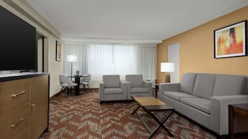 Quarto de hotel com sofá, duas poltronas, mesa de centro, televisão, escrivaninha com cadeira e tapete estampado.