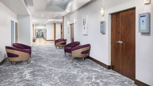 A hotel corridor with a marble floor, purple and beige armchairs, wooden doors, and wall-mounted light fixtures.