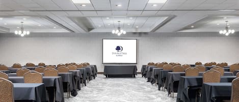 Un salón de conferencias con filas de mesas y sillas, una pantalla de proyector mostrando un logotipo, y piso alfombrado.