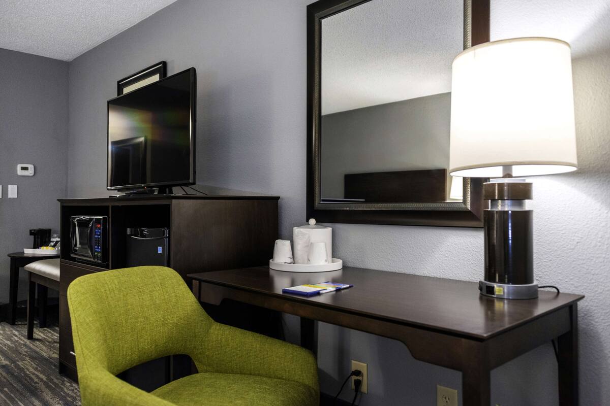 A hotel room with a desk, a green chair, a television, a lamp, and a mirror.