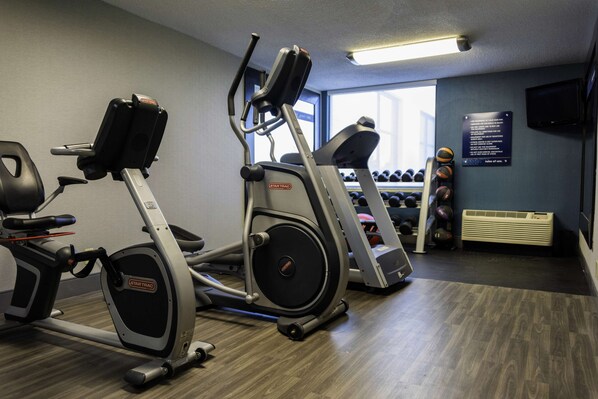 A gym with two elliptical machines, a set of dumbbells, and a wall-mounted TV.