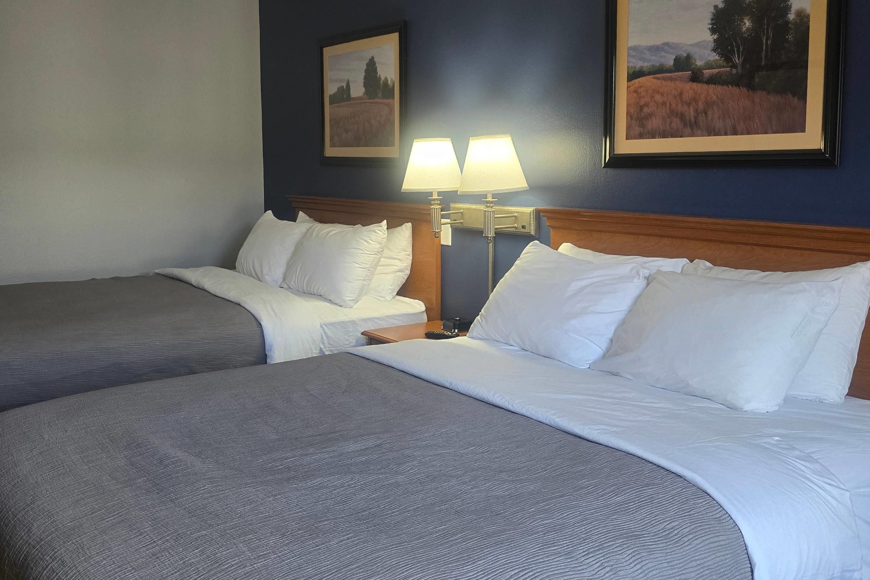 A hotel room with two beds, a nightstand with lamps, and two framed landscape paintings on the wall.