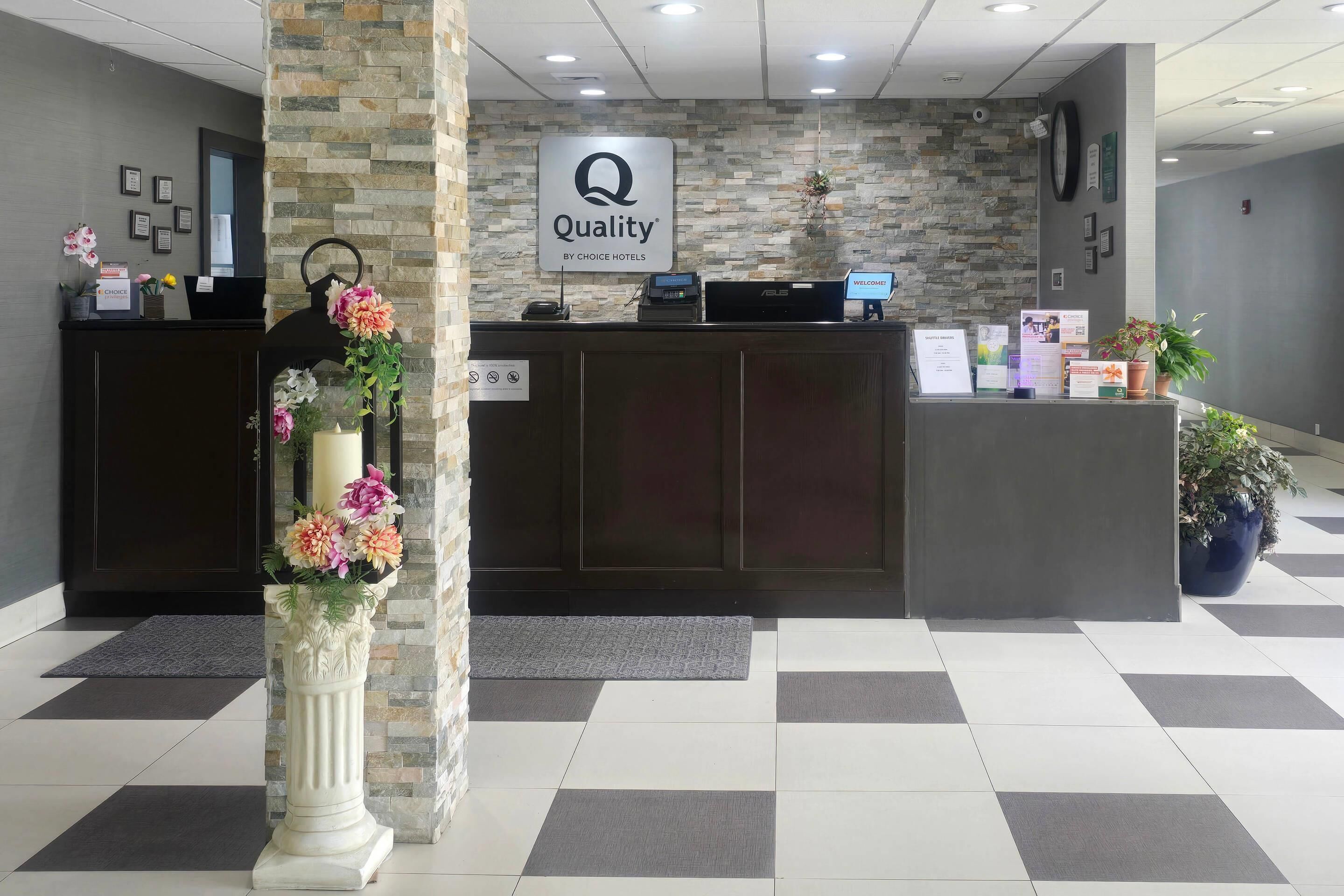 Uma área de recepção de hotel com um fundo de parede de pedra, um balcão de recepção, um quiosque digital e um vaso de planta.