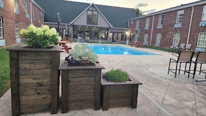A courtyard with a swimming pool, wooden planters with plants, outdoor seating, and a brick building with large windows.