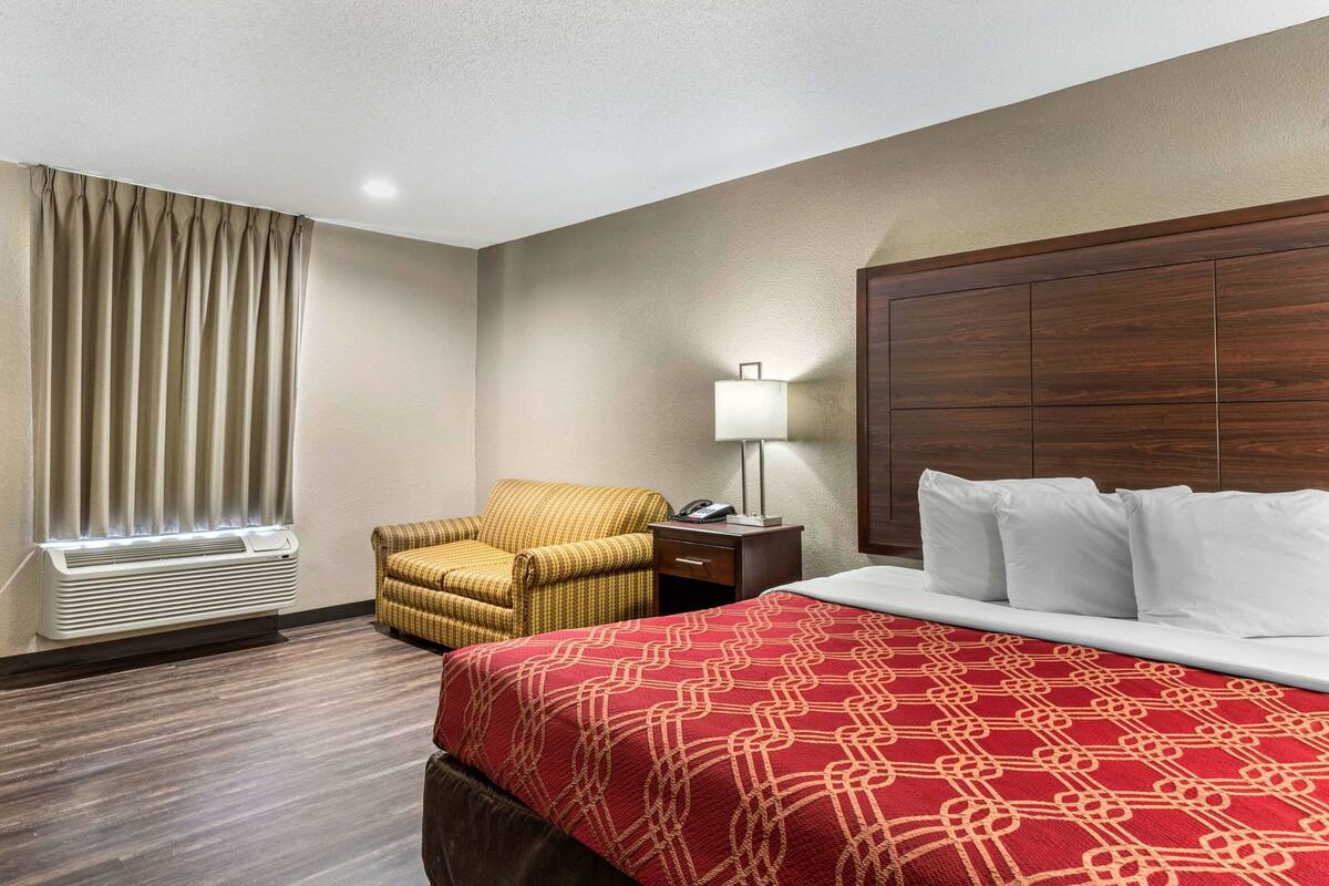 Quarto de hotel com cama, uma cadeira, abajur e janela com cortinas.
