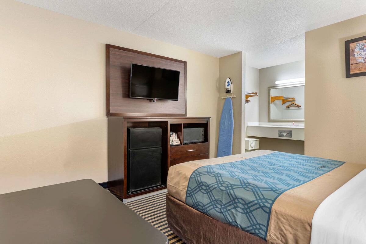 Quarto de hotel com cama, escrivaninha, televisão na parede, mini-geladeira e espelho com porta-toalhas.