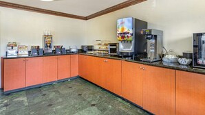 Une cuisine d’hôtel avec des meubles de rangement orange, une machine à café, un distributeur de jus et un présentoir à pain.