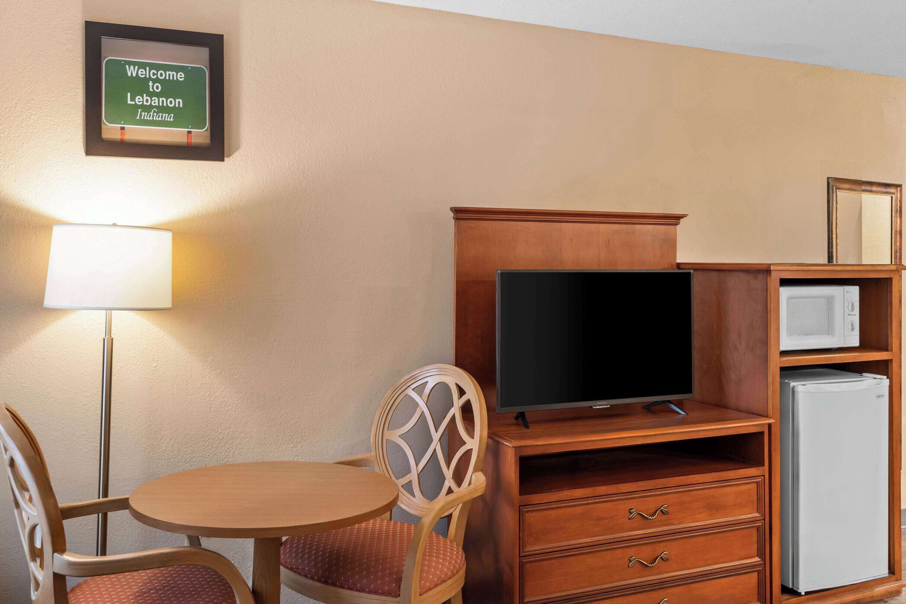 A hotel room with a wooden desk, a television, a refrigerator, a chair, and a lamp.