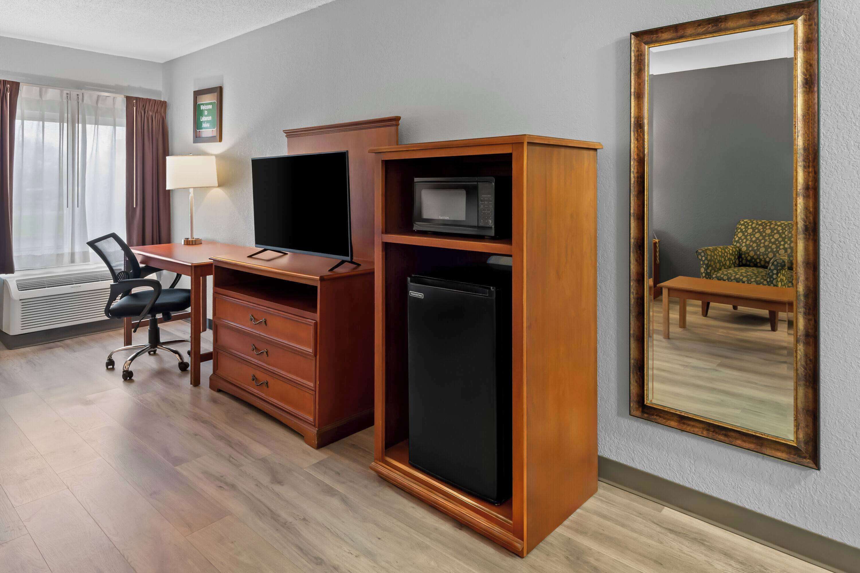 Una habitación de hotel con un escritorio de madera, un refrigerador negro, un televisor, una lámpara, una silla y un espejo grande en la pared.