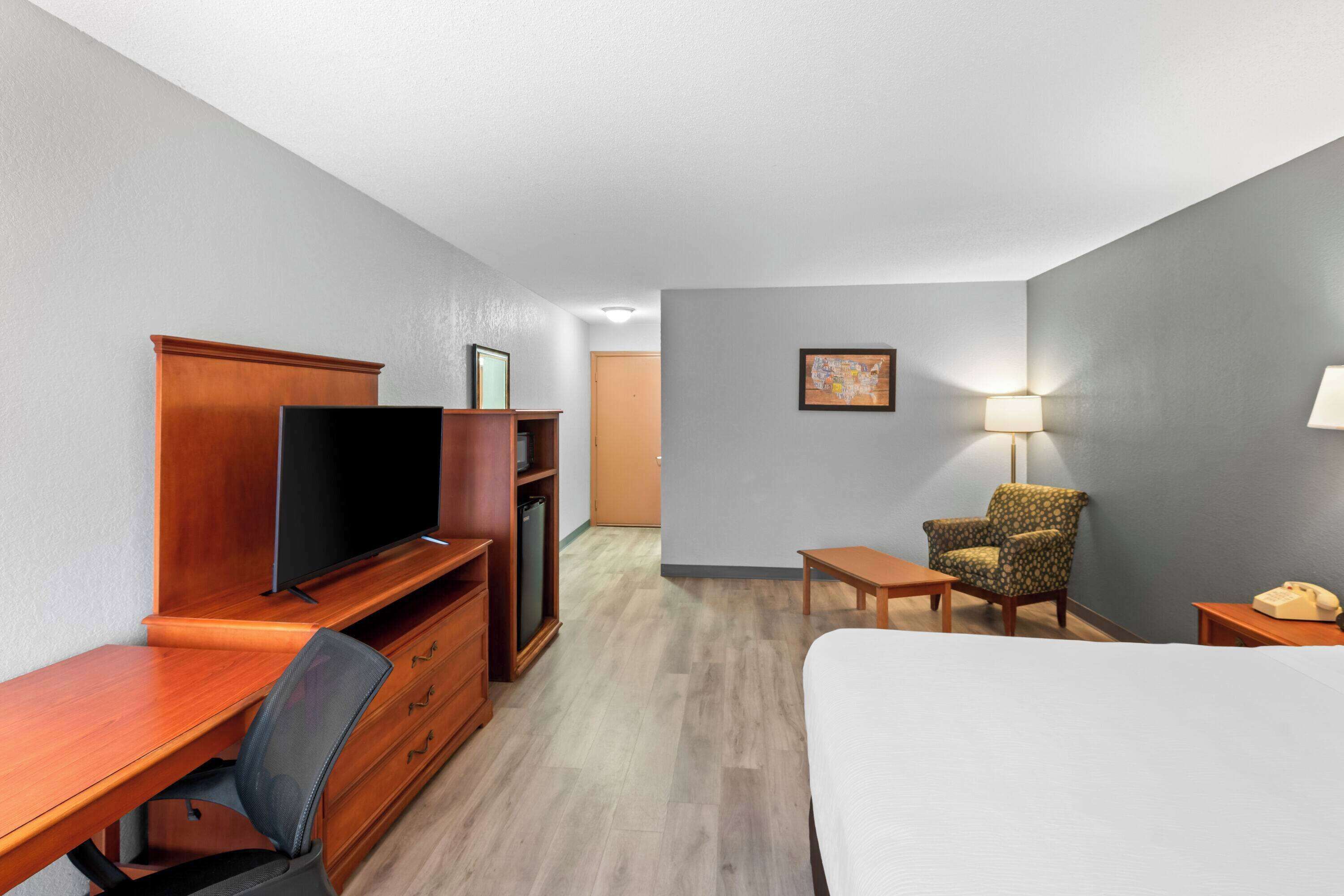 Quarto de hotel com uma escrivaninha de madeira, televisão, cama, cadeira, mesa pequena e luminária.