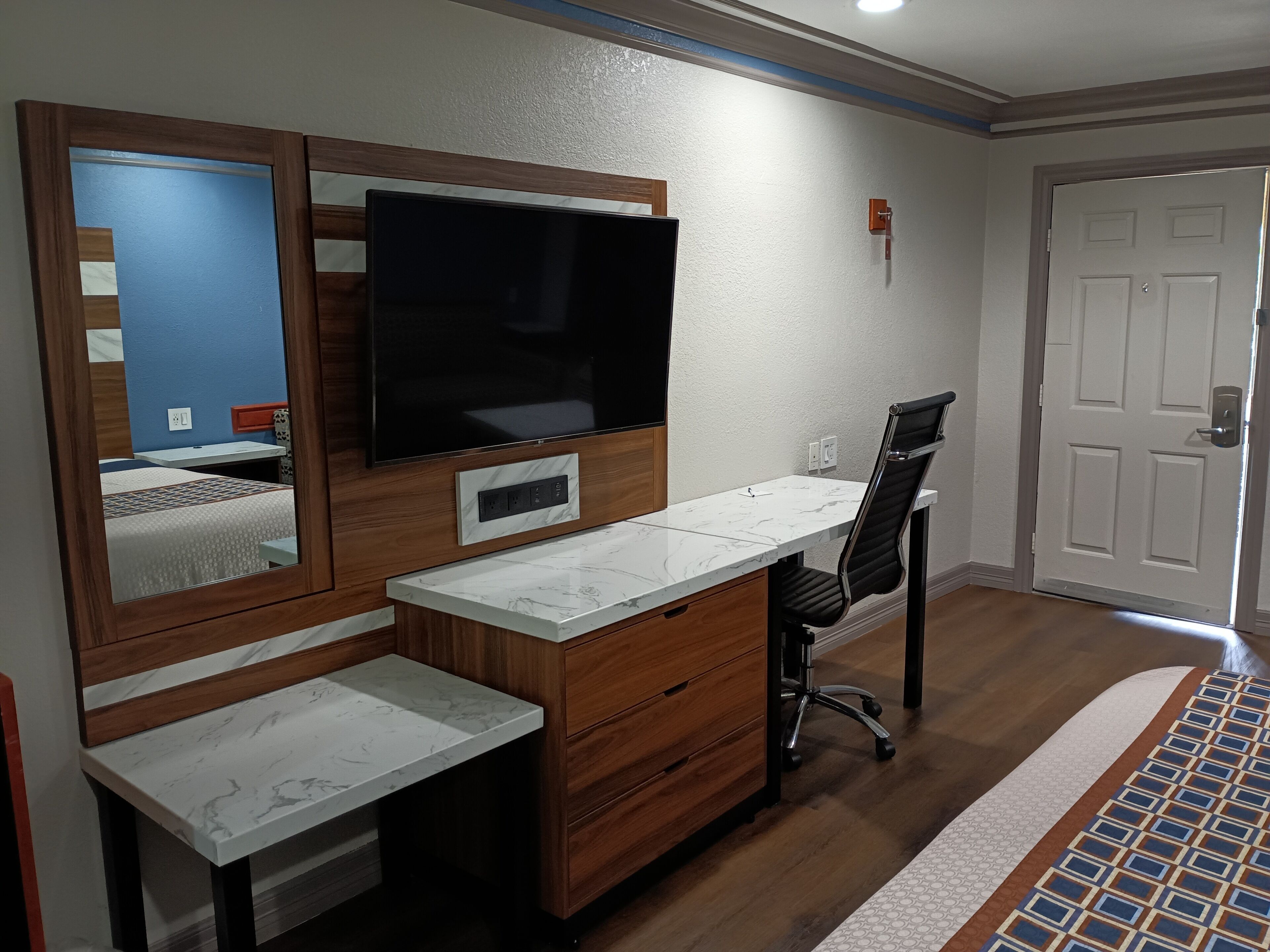 A hotel room with a large flat-screen TV mounted on the wall, a wooden desk with drawers, a black office chair, and a white door.