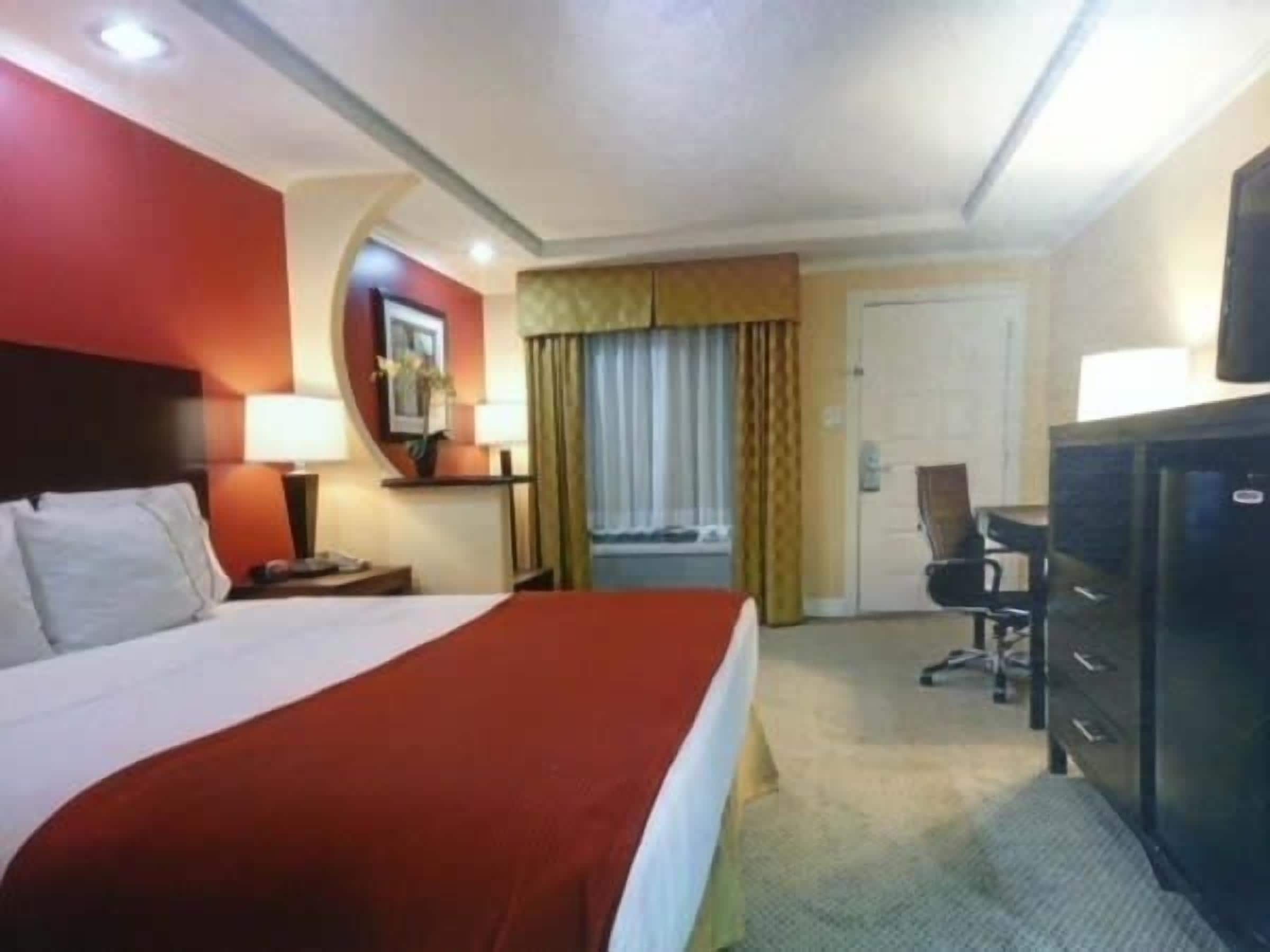 A hotel room with a large bed, a desk with a lamp, a television, and a wardrobe.