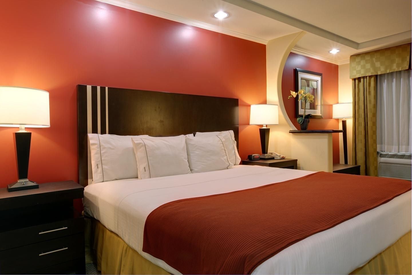 Habitación de hotel con una cama grande, dos mesitas de noche con lámparas, una mesita auxiliar, un televisor montado en la pared y una ventana con cortinas.