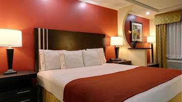 Habitación de hotel con una cama grande, dos mesitas de noche con lámparas, una mesita auxiliar, un televisor montado en la pared y una ventana con cortinas.