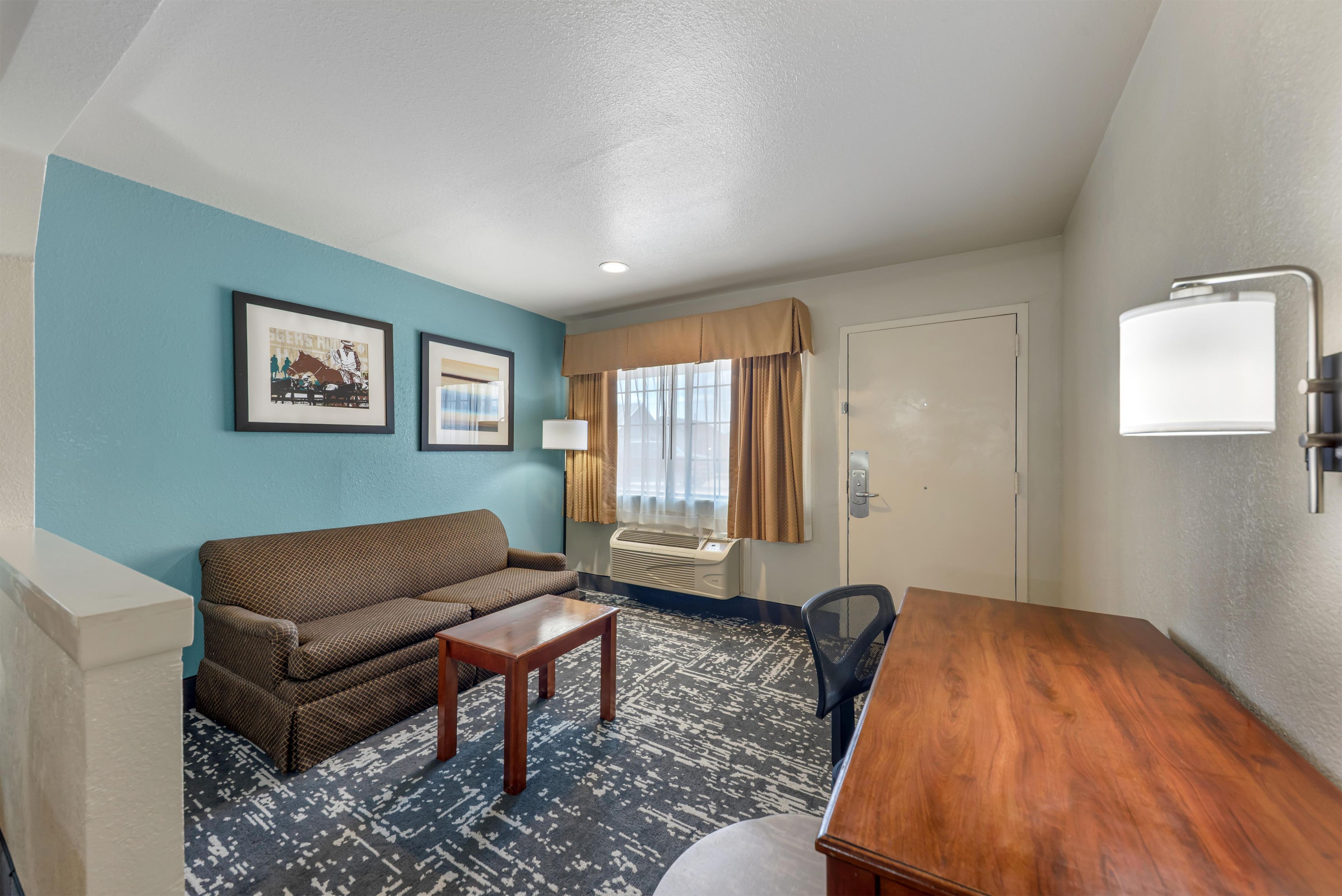 Une chambre d’hôtel avec un canapé, une petite table, un bureau, une lampe et une fenêtre avec des rideaux.