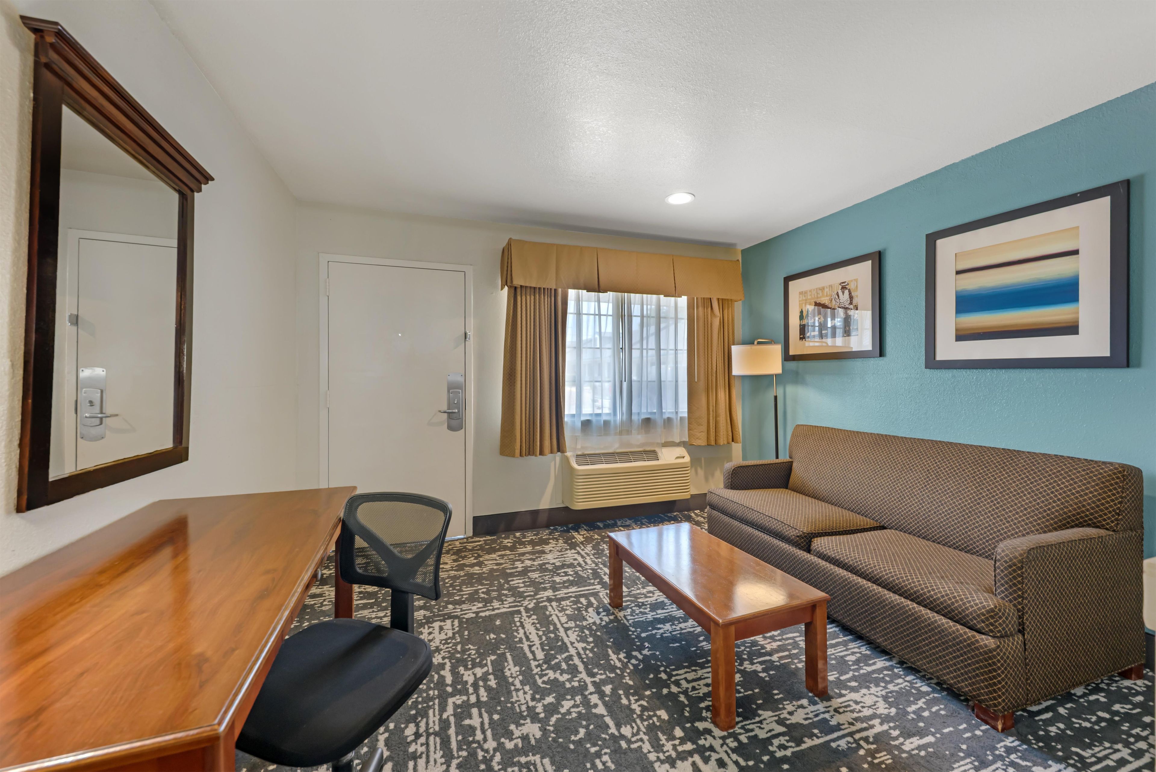 Une chambre d’hôtel avec un bureau en bois, un fauteuil de bureau noir, un canapé marron, un tapis à motifs, une fenêtre avec des rideaux et deux tableaux encadrés accrochés au mur.