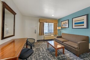 A hotel room with a wooden desk, a black office chair, a brown sofa, a patterned rug, a window with curtains, and two framed pictures on the wall. - Best Western Regency Inn & Suites (Gonzales)