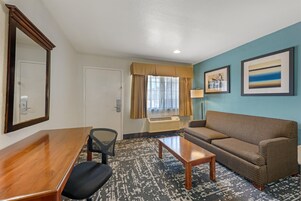 Habitación de hotel con un escritorio de madera, una silla de oficina negra, un sofá marrón, un tapete estampado, una ventana con cortinas y dos cuadros en la pared.