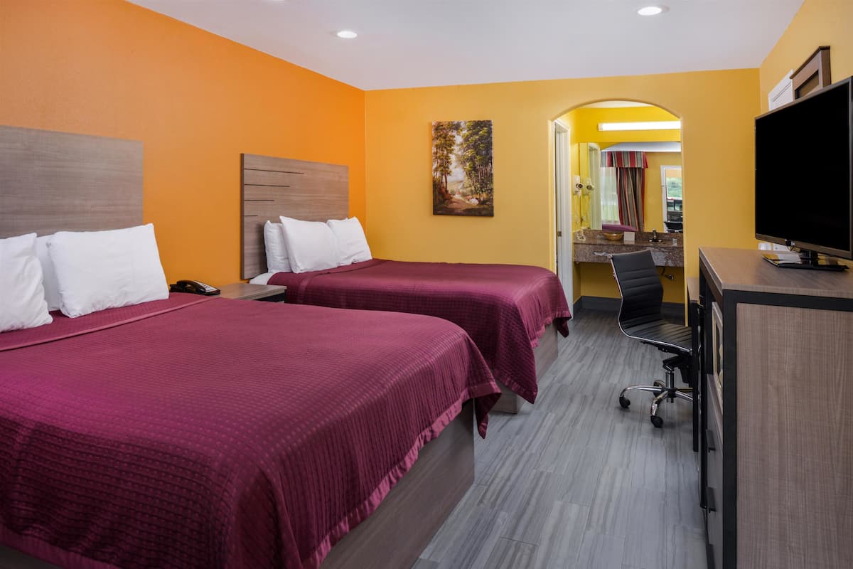 A hotel room with two beds, a desk, a TV, and a bathroom visible through an archway.