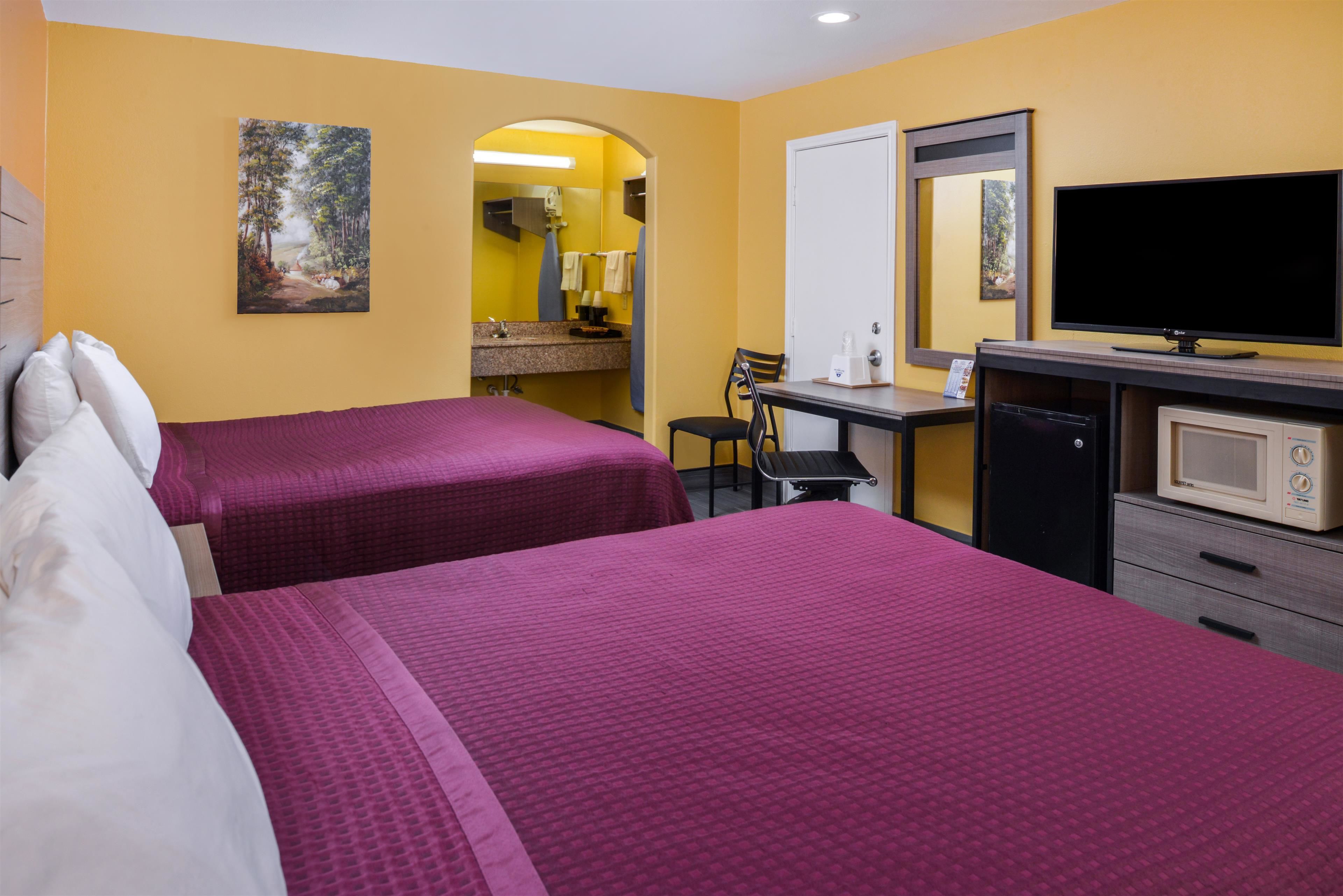 Habitación de hotel con dos camas, un escritorio con silla, televisión, microondas y un baño visible a través de un arco.