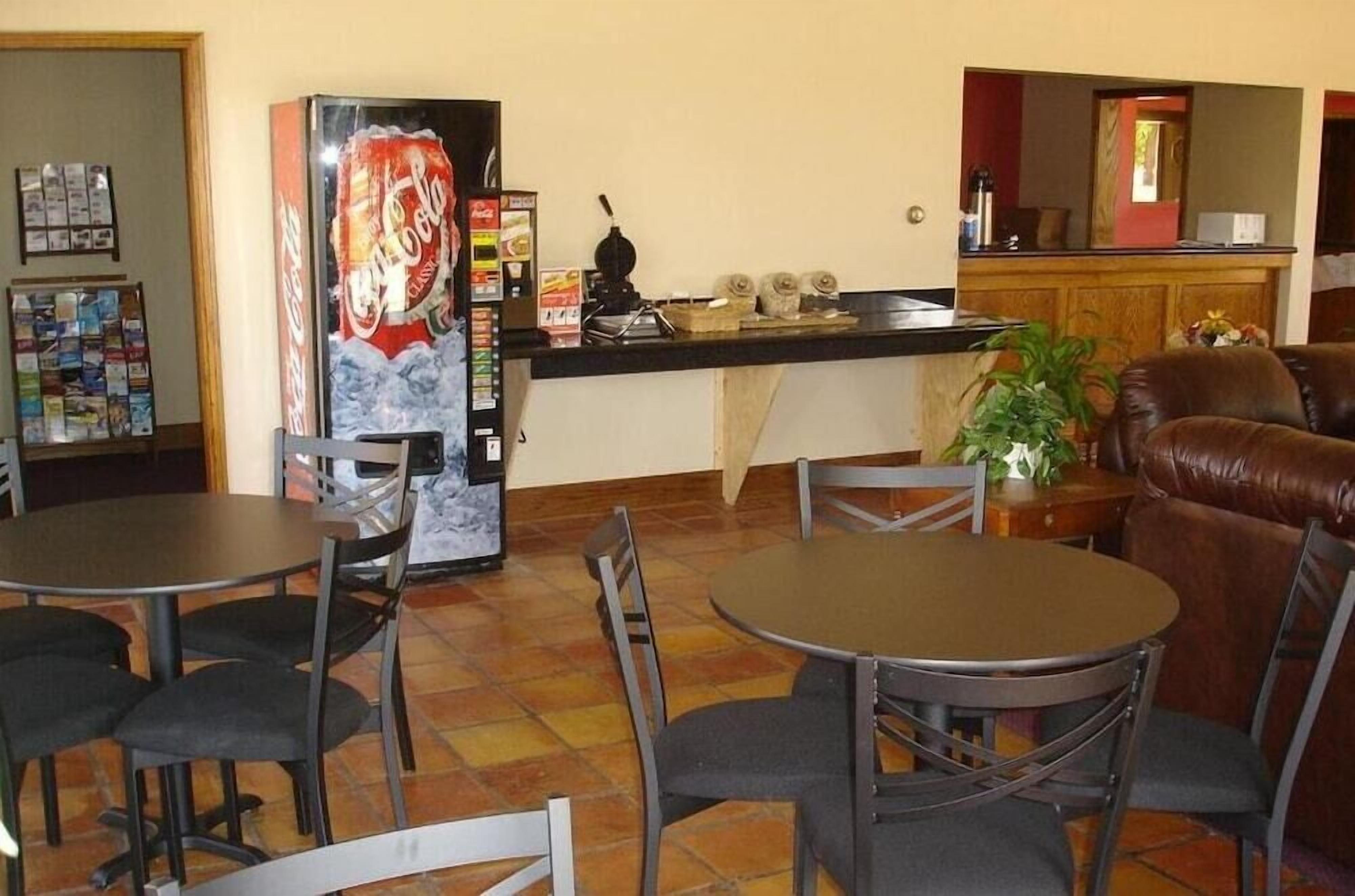 A dining area with tables and chairs, a refrigerator with a Coca-Cola logo, a counter with a coffee machine, and a leather couch.