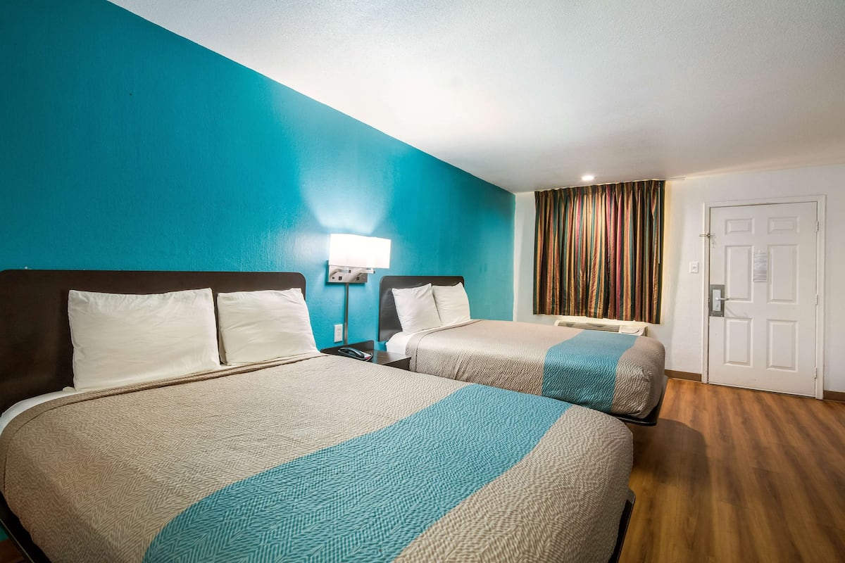 Habitación de hotel con dos camas, una pared de acento color aguamarina, piso de madera, una mesita de noche con lámpara y una puerta con cortina.