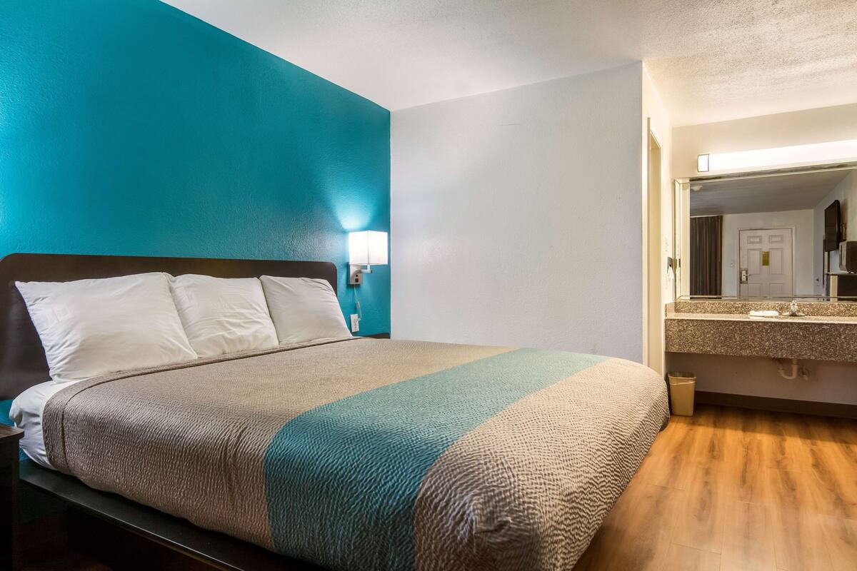 Habitación de hotel con cama, mesita de noche, baño con lavamanos y espejo, y paredes en tonos aguamarina y blanco.