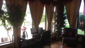 A room with a desk, computer, chair, and large windows with curtains. There are potted plants and a view of greenery outside.