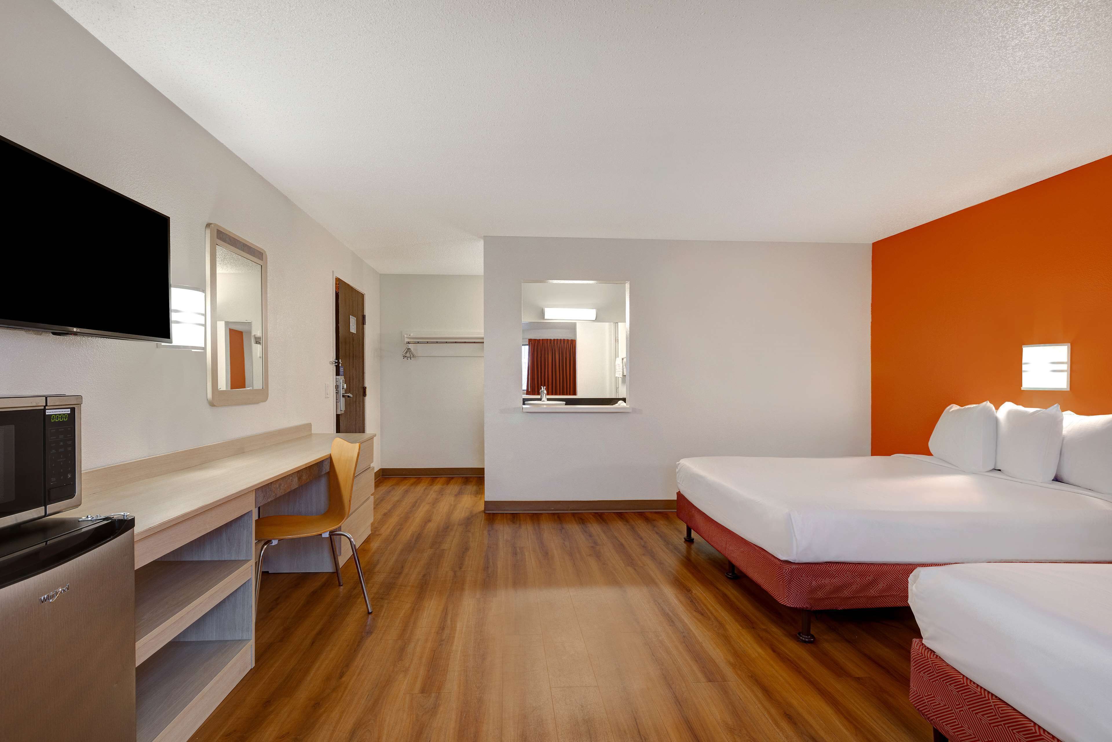 Habitación de hotel con piso de madera, una cama, un escritorio con televisor y un espejo.