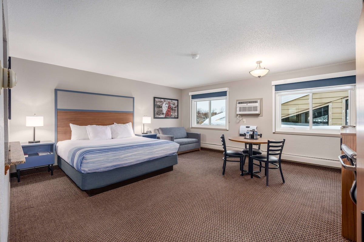 A hotel room with a bed, a desk with chairs, a small table, a sofa, and a window with blinds.