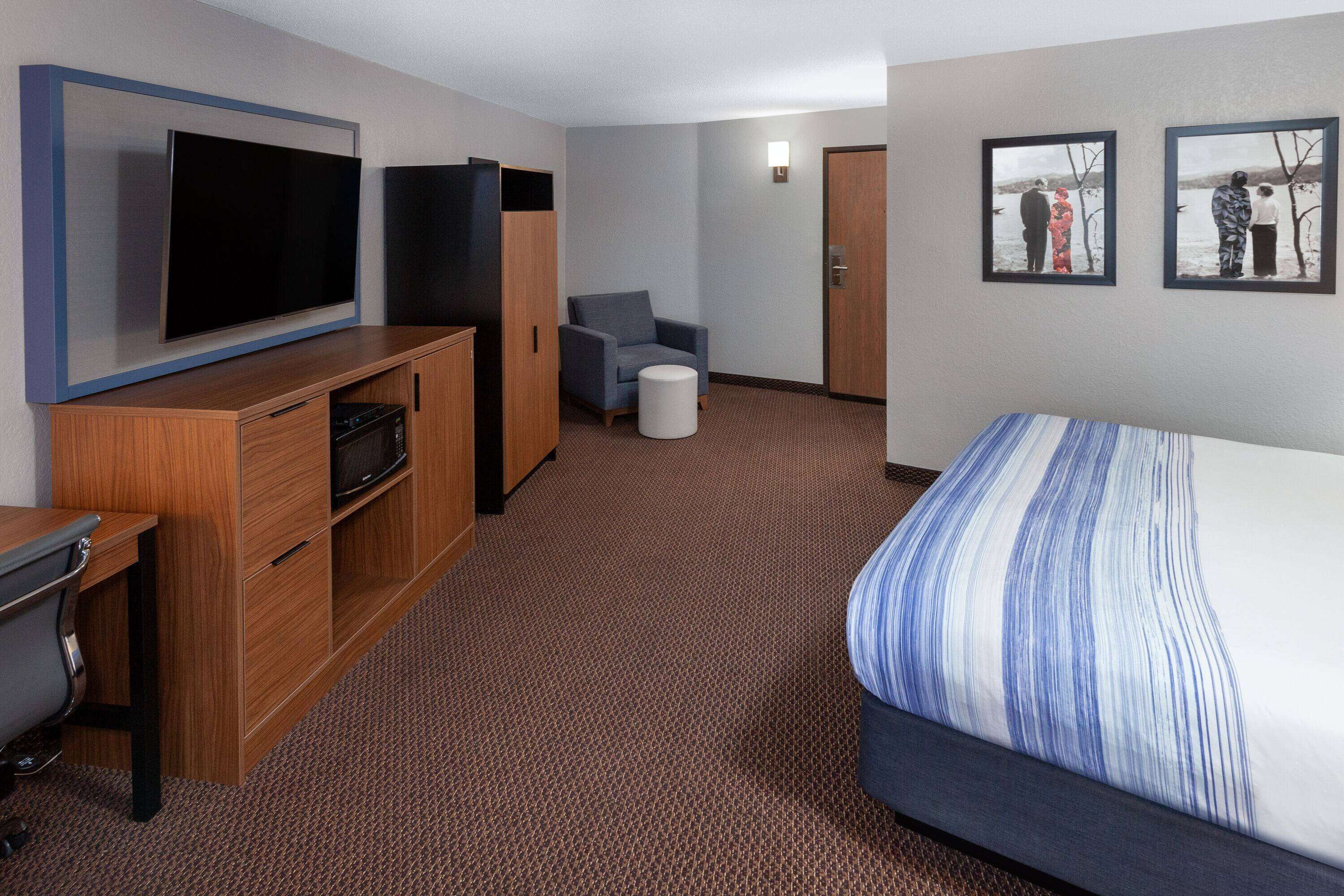 A hotel room with a large bed, a flat-screen TV mounted on the wall, a desk with a chair, and a small seating area with a chair and ottoman.