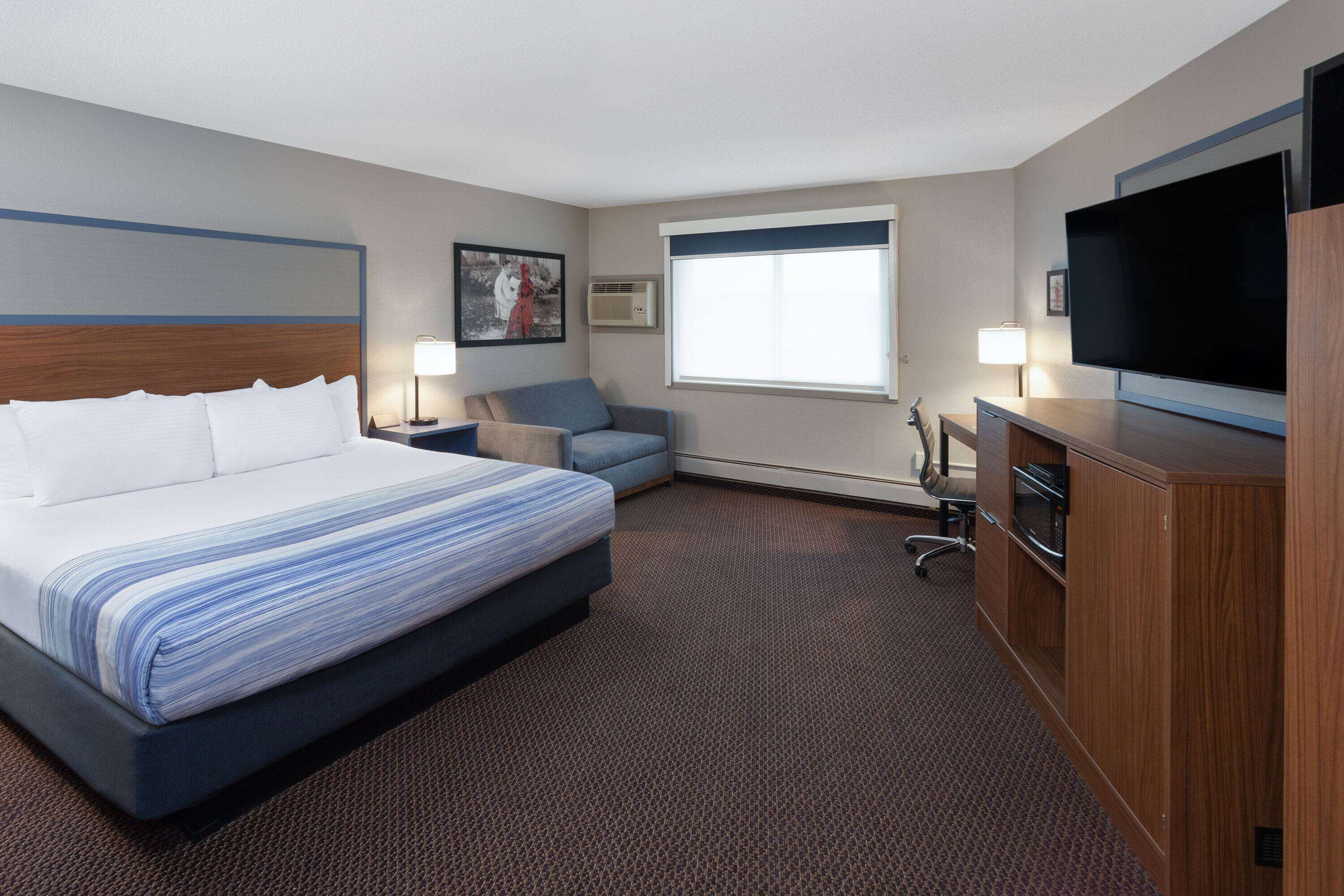 A hotel room with a large bed, a desk with a flat-screen TV, a chair, a desk lamp, and a window with blinds.