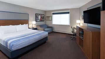 A hotel room with a large bed, a desk with a flat-screen TV, a chair, a desk lamp, and a window with blinds.
