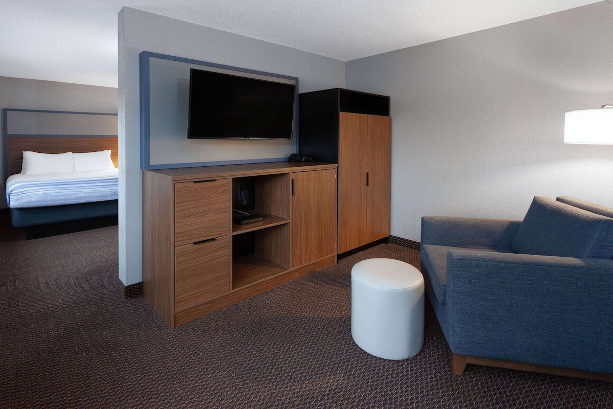 A hotel room with a bed, a TV mounted on a wooden cabinet, a sofa, and a small white ottoman.