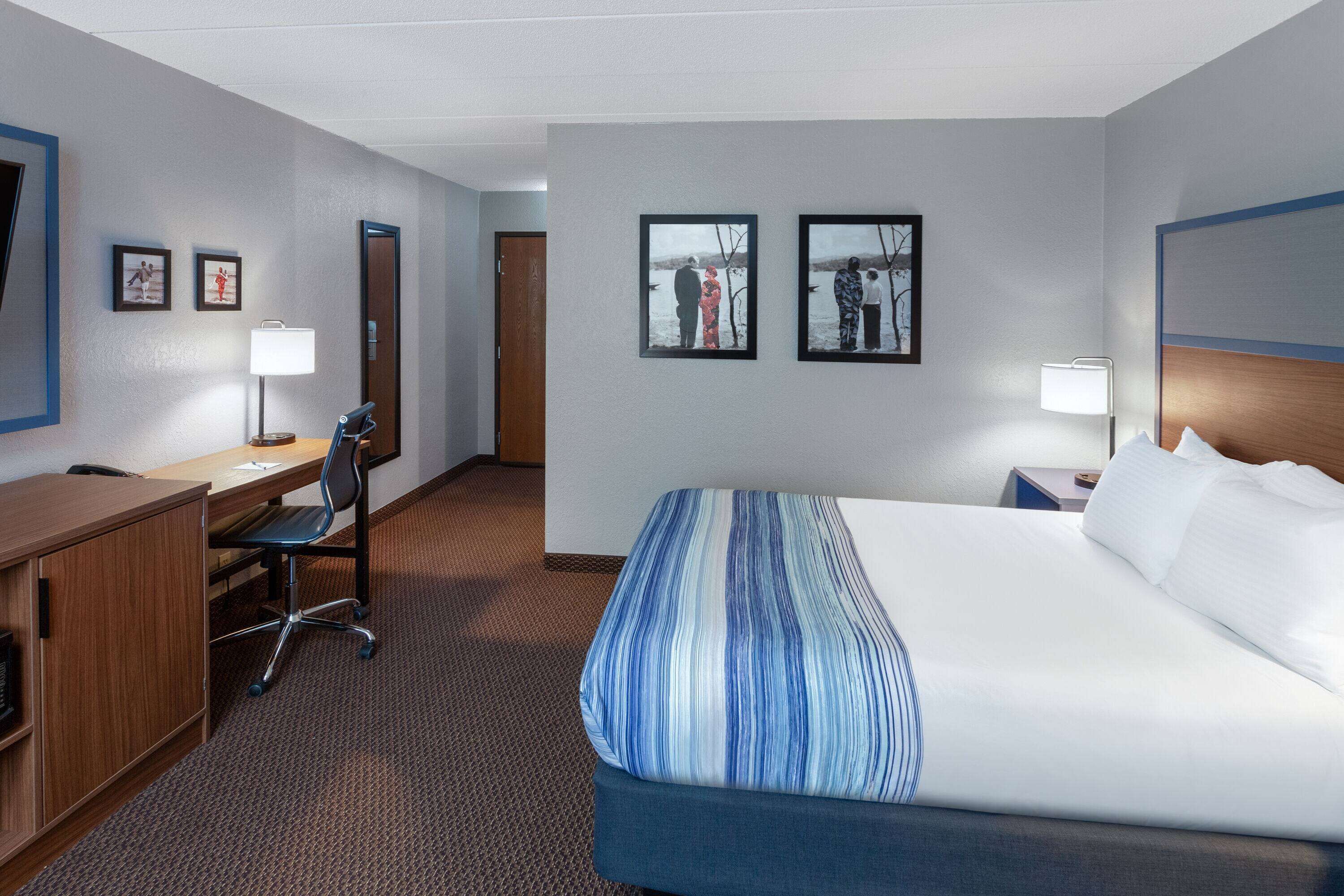A hotel room with a bed, desk, chair, and two framed pictures on the wall.