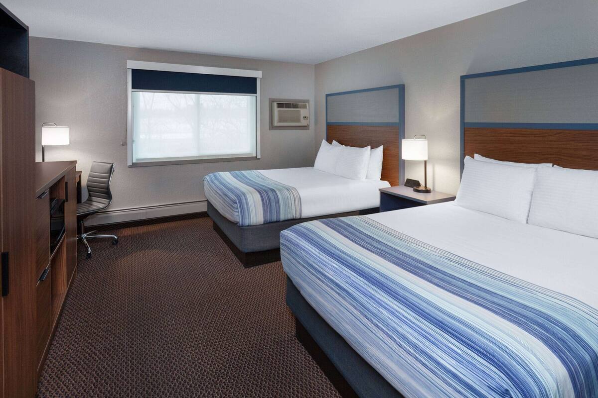 A hotel room with two beds, a desk, a chair, a window, and a wall-mounted air conditioner.