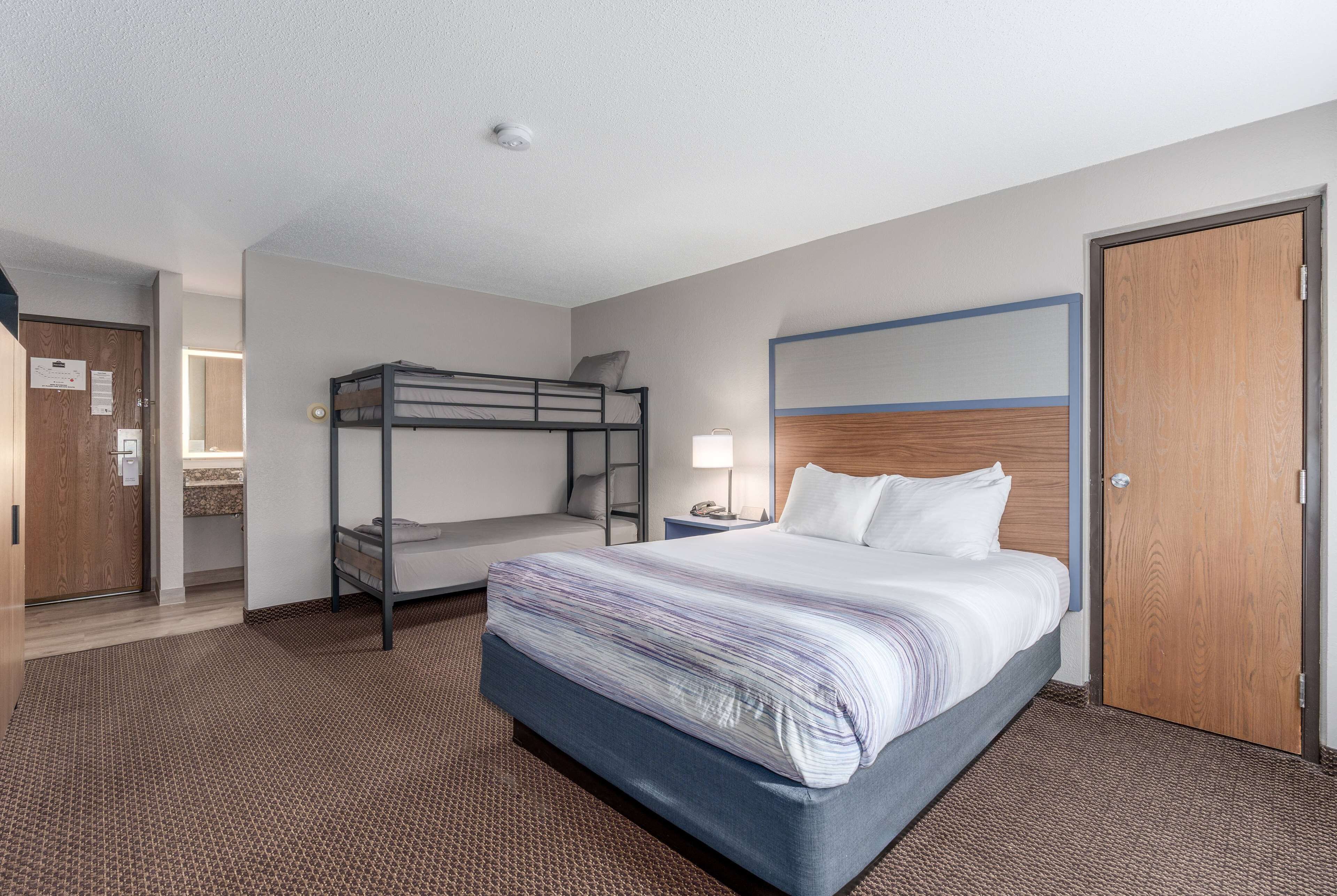 Ein Hotelzimmer mit Etagenbett, einem Einzelbett, einer Holztür, einem Nachttisch mit Lampe und einem gemusterten Teppich.