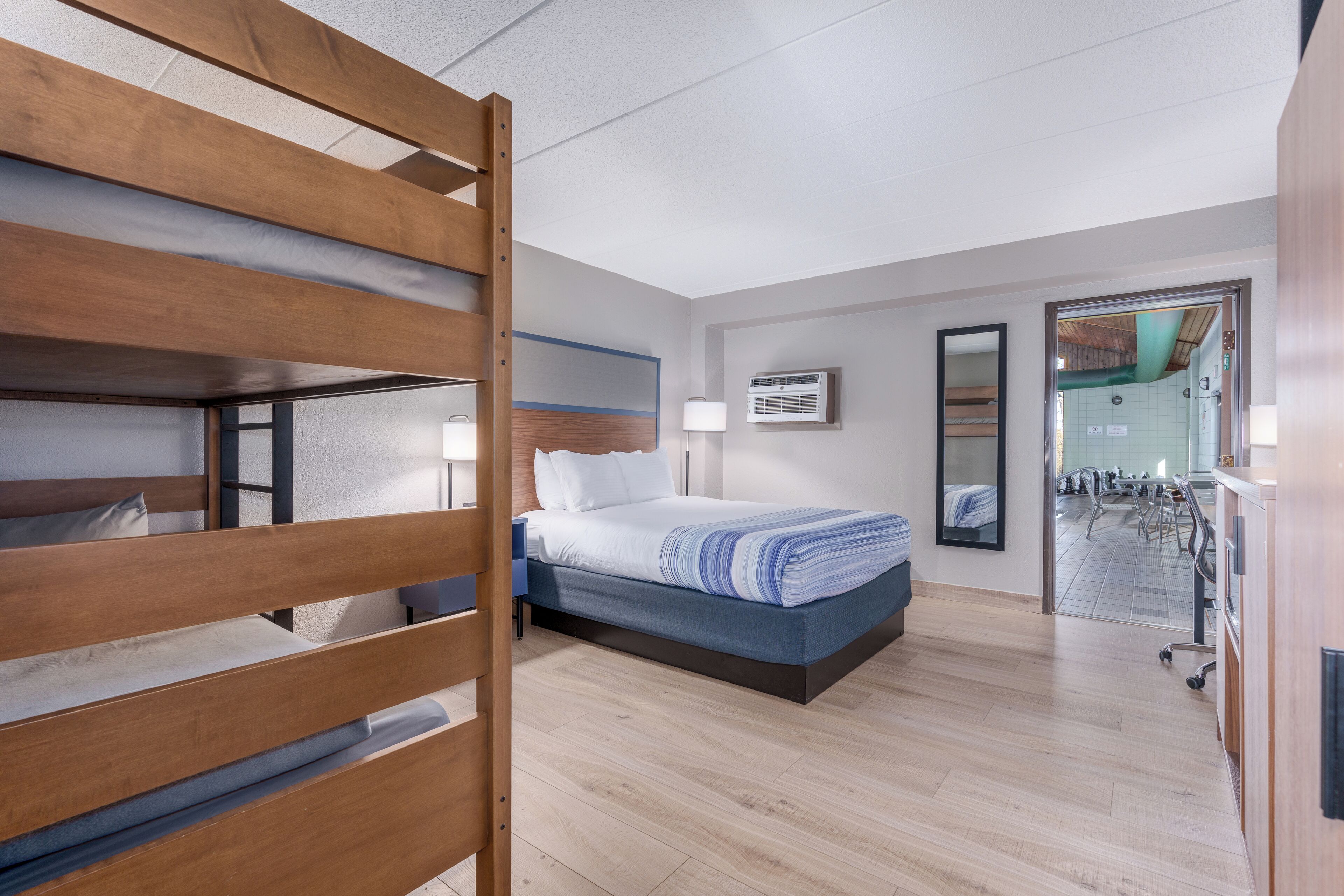 A modern hotel room with a bunk bed, a single bed, a desk, and a bathroom visible through an open door.