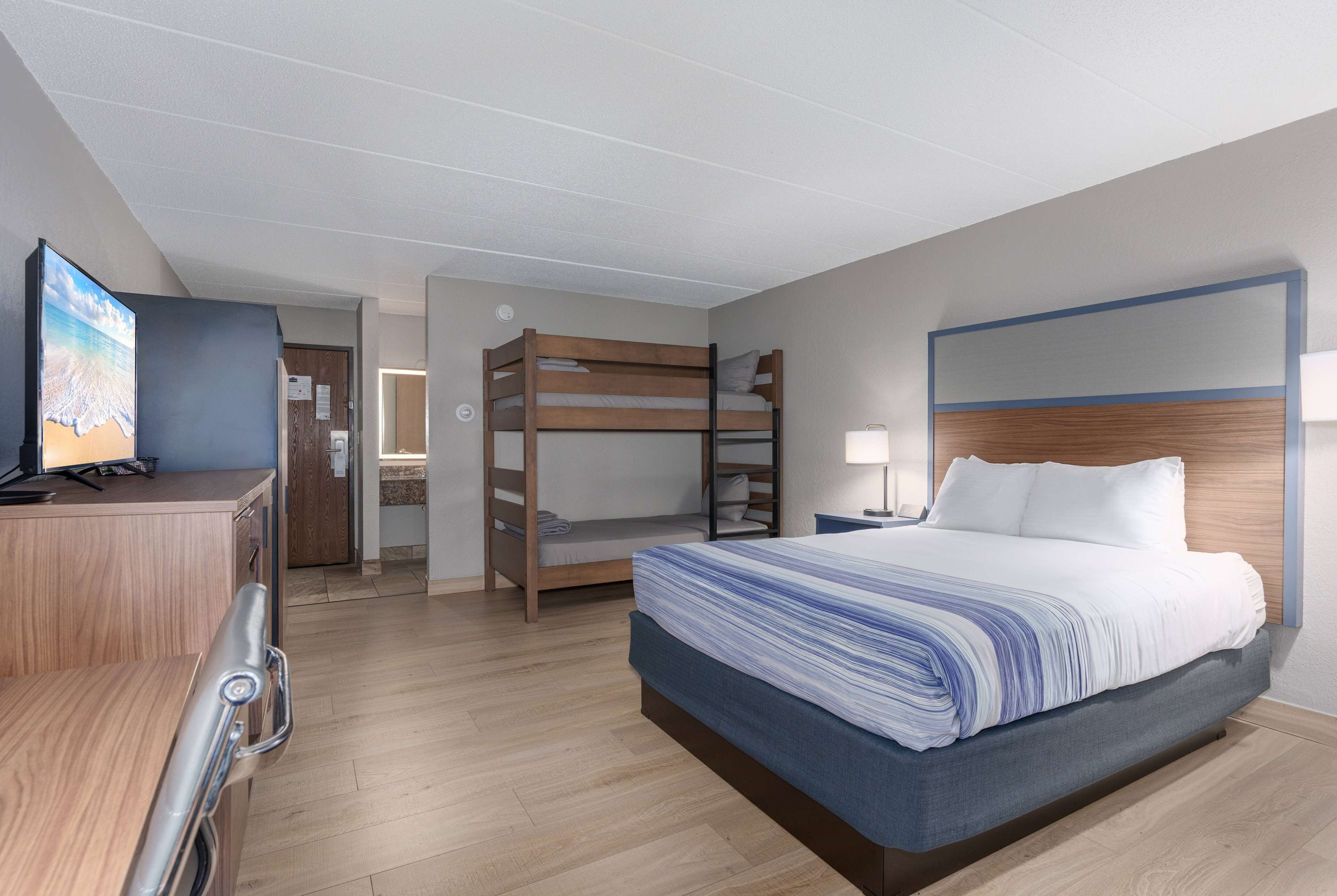 A hotel room with a bunk bed, a desk with a computer, a TV, and a bedside table with a lamp.
