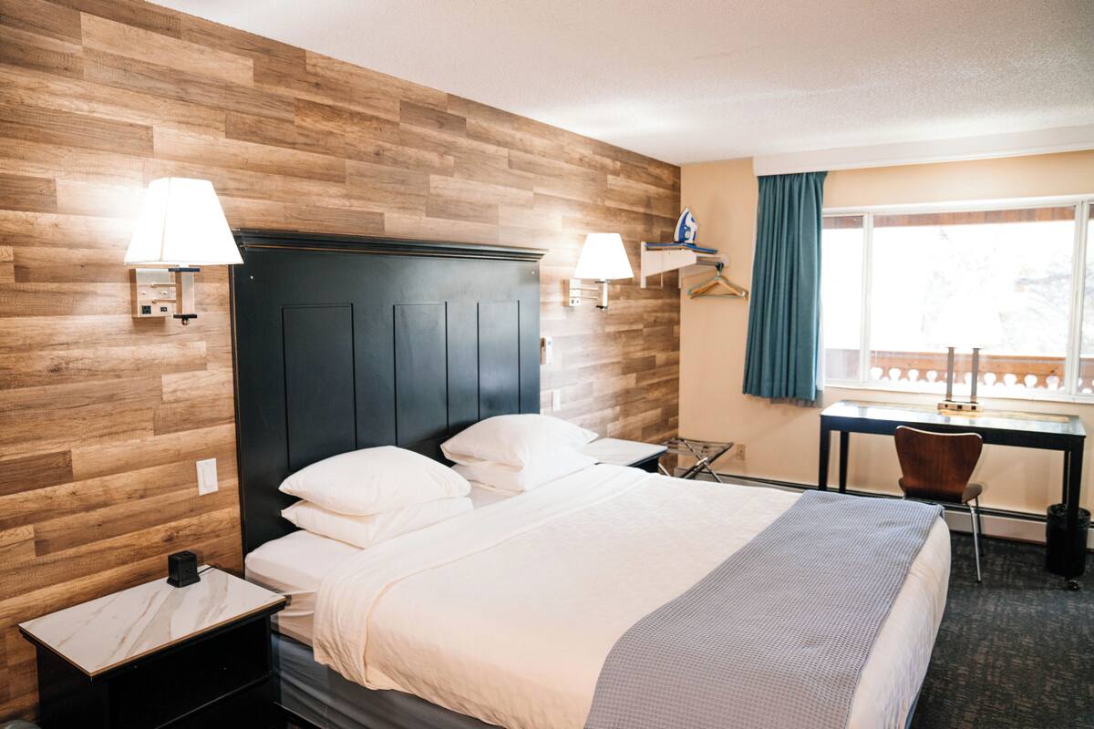 A hotel room with a wooden wall, a bed with white linens, two bedside lamps, a desk with a chair, and a window with a curtain.