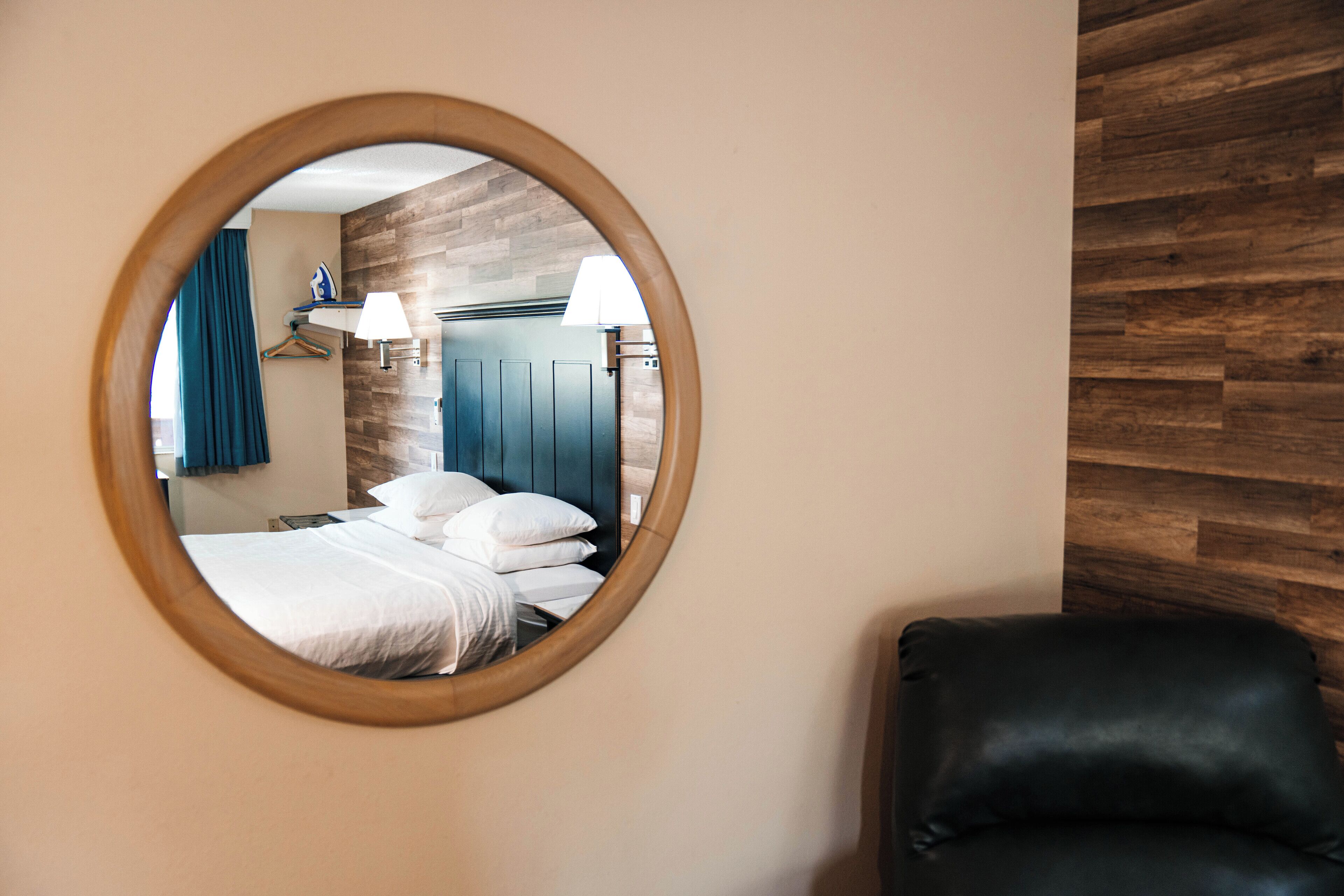 Un espejo circular que refleja un dormitorio con cama, almohadas, una prenda azul colgada y una lámpara fijada a la pared.