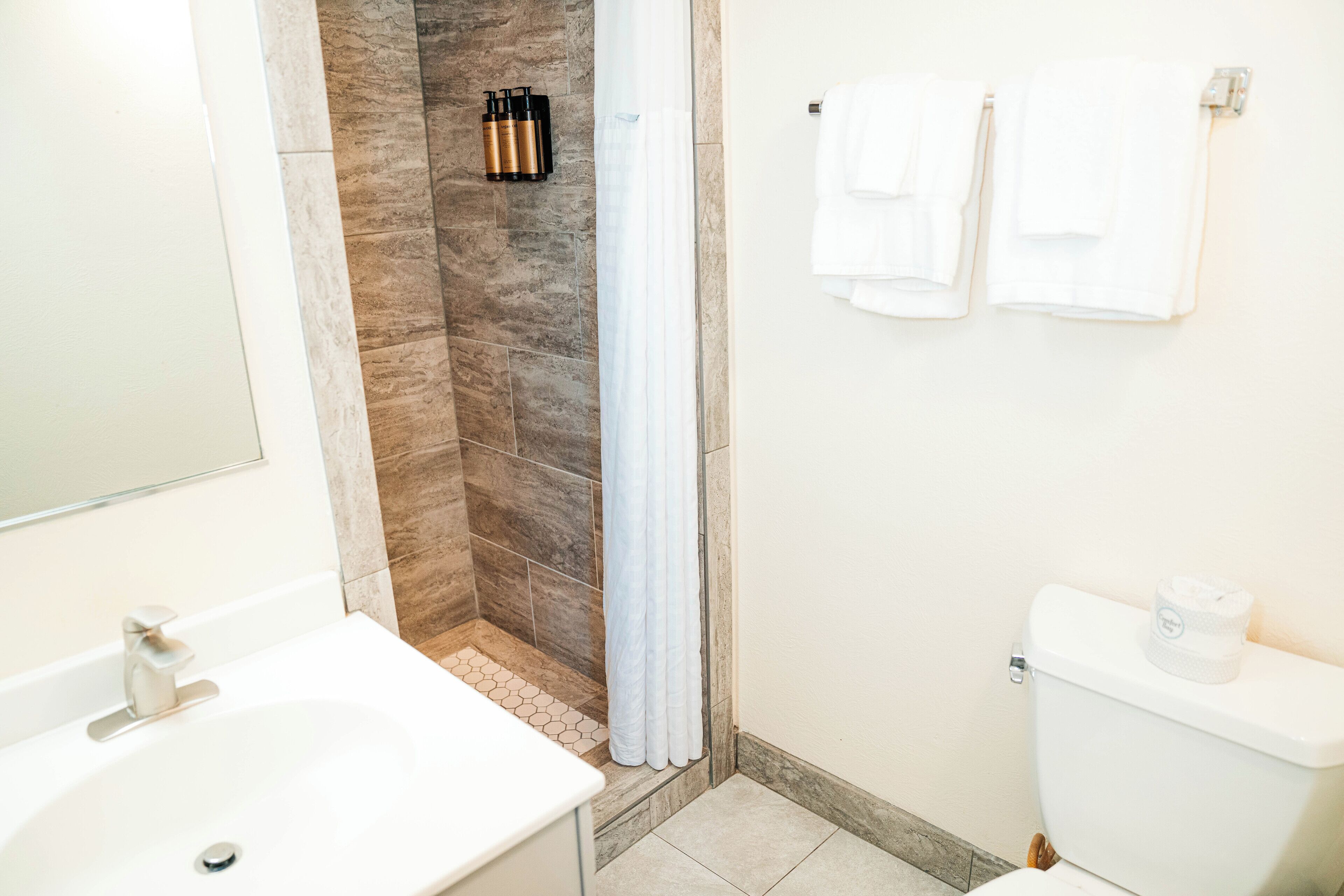 Une salle de bain avec un lavabo blanc, une douche avec un mur en pierre et des toilettes sur lesquelles est posé un rouleau de papier toilette.