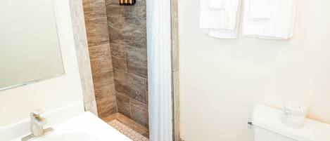 Une salle de bain avec un lavabo blanc, une douche avec un mur en pierre et des toilettes sur lesquelles est posé un rouleau de papier toilette.