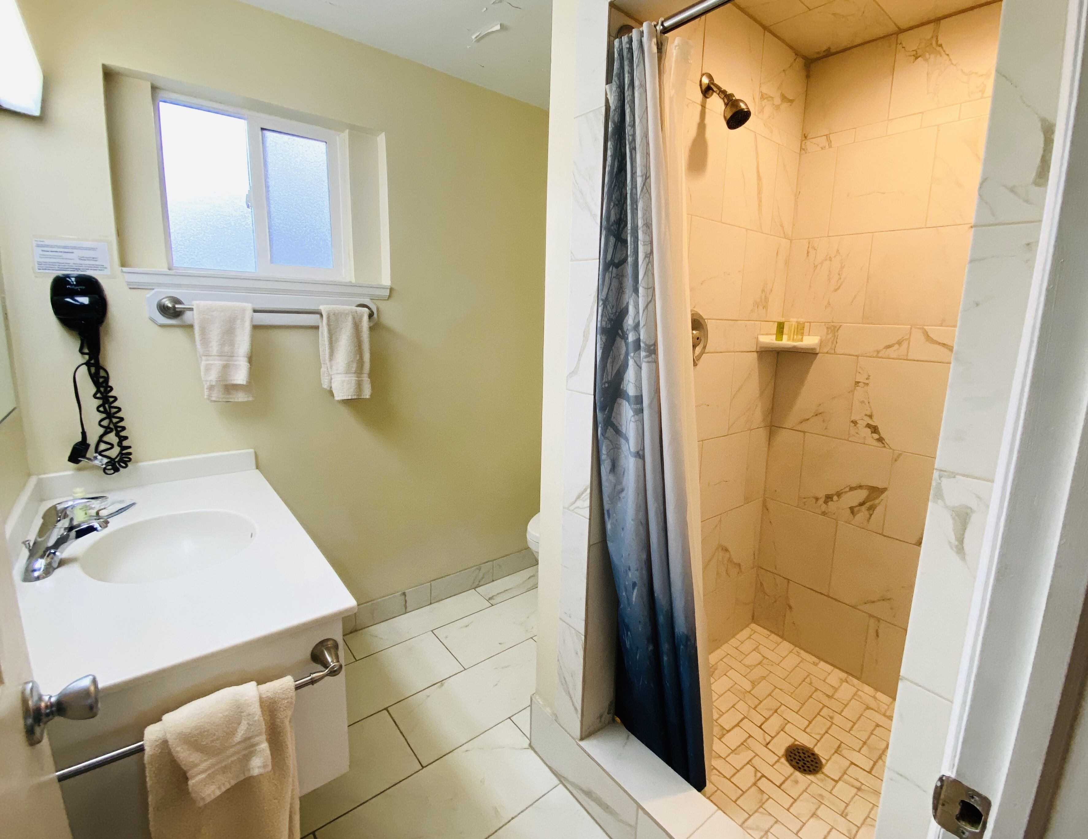 Un baño con lavamanos, secador de pelo, ventana y ducha con cortina.