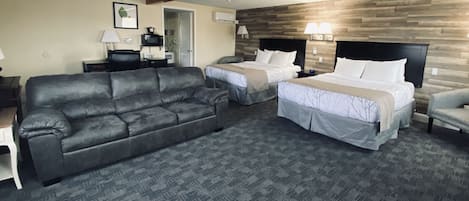 A hotel room with a gray sofa, two beds with white linens, a wooden accent wall, and a gray carpet.