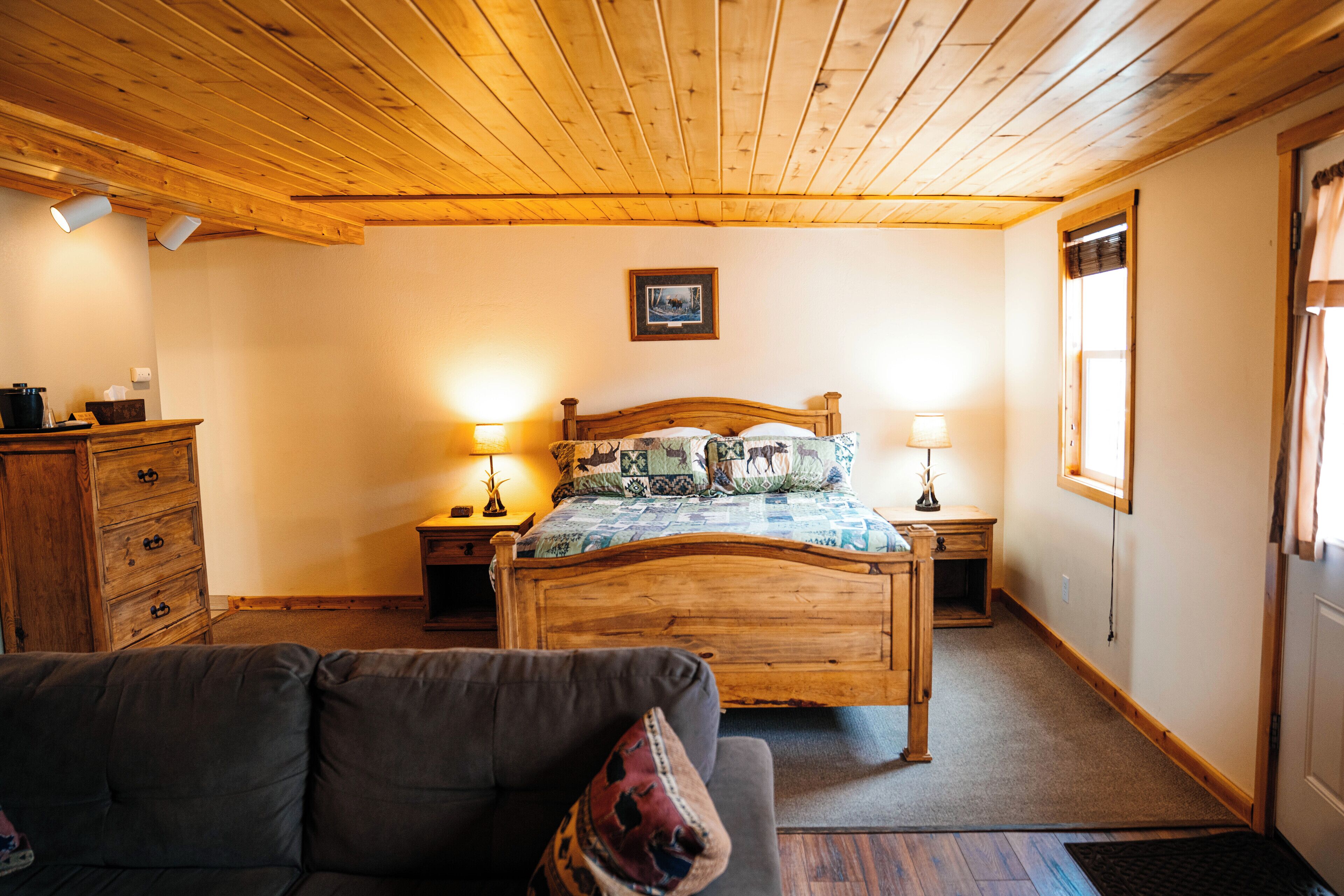 A cozy bedroom with a wooden ceiling, a bed with patterned bedding, two bedside lamps, a wooden dresser, and a view of the outdoors through a window.
