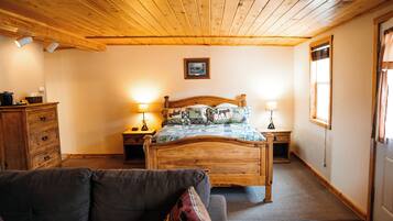 A cozy bedroom with a wooden ceiling, a bed with patterned bedding, two bedside lamps, a wooden dresser, and a view of the outdoors through a window.