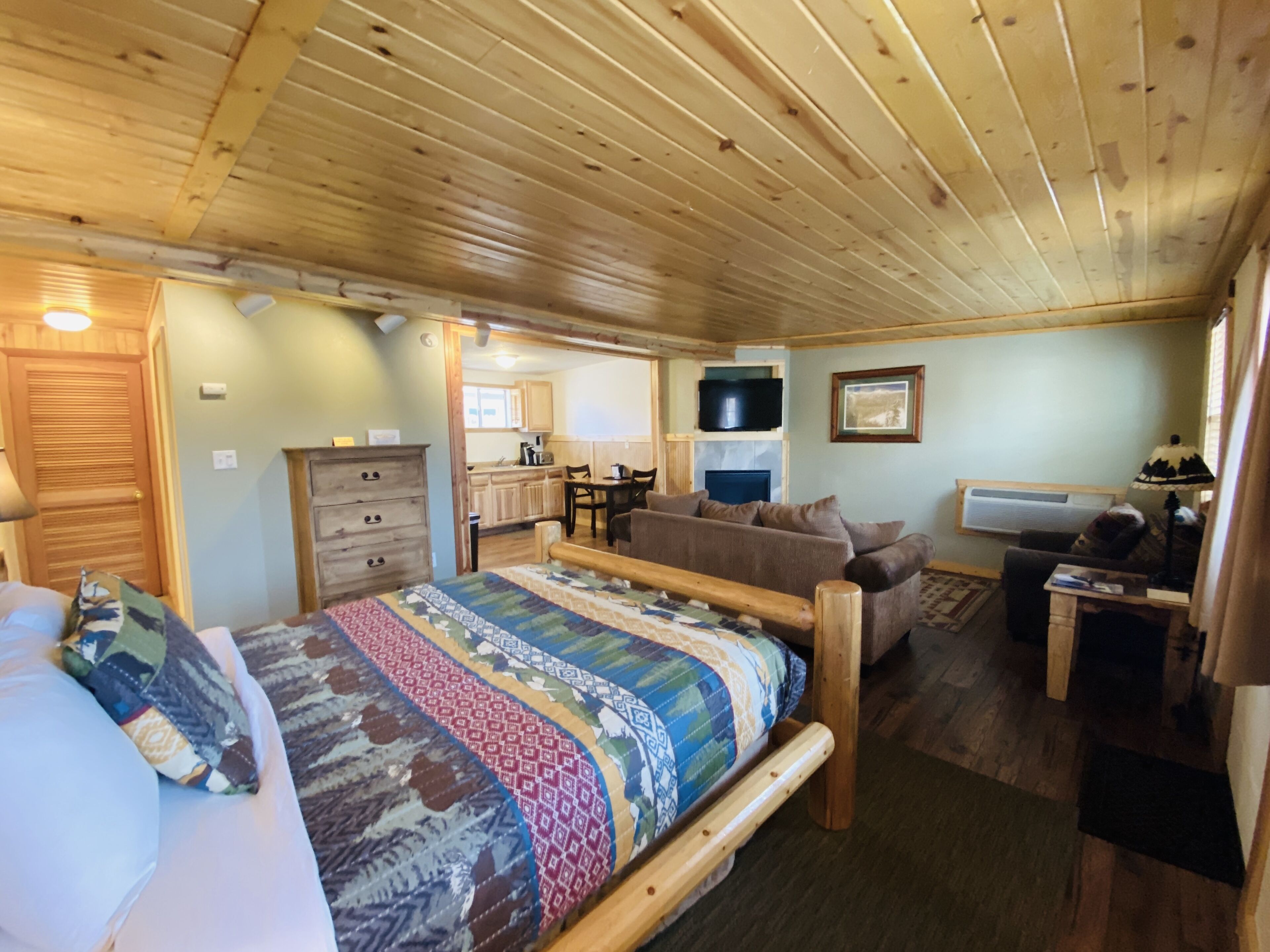 Een knus slaapkamer in chaletstijl met een houten plafond, een groot bed met een kleurrijke quilt, een bank, een kleine tafel en een tv aan de muur bevestigd.